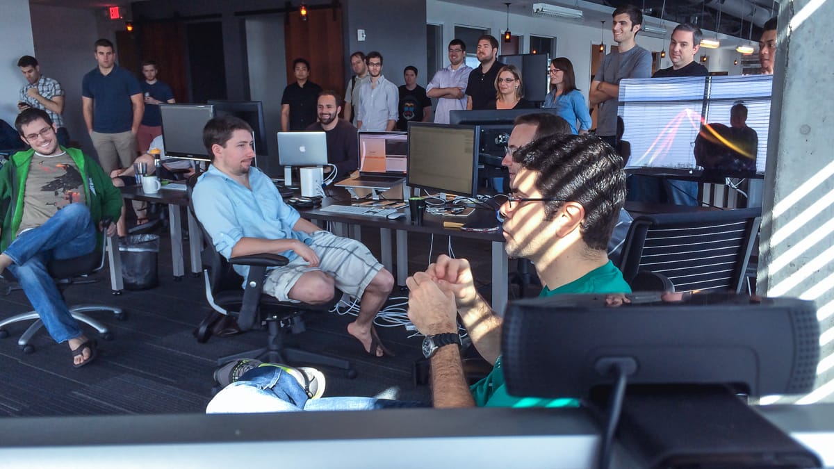 Picture of more than twenty people crowded together in the back of an office near an iMac sporting a tiny webcam in the foreground.